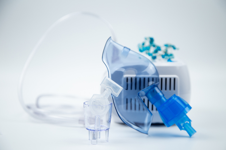 Close-up of an inhalation bowl with an ampoule of medicinal solution in it against the background of a blue breathing mask and a device on a white background. The concept of treatment of respiratory tract diseases, lung diseases, medicines.