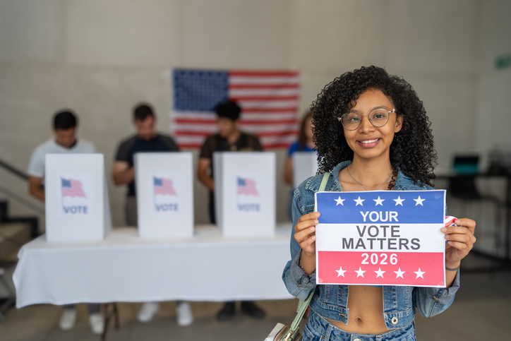 African American woman smiles holding a 'Your Vote Matters 2026' sign at a US election polling station. In the background, a diverse group of people votes in voting booths, democracy in America
