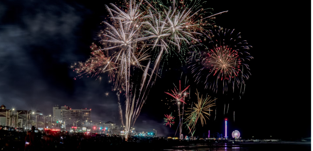 Vibrant fireworks display over a coastal city skyline at night, with a ferris wheel and other amusement park rides visible.